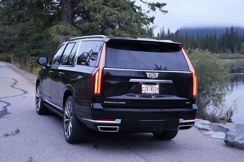The Escalade seen from behind on a lakeside road, full-height tail lamps lit, forest on one side and water on the other.