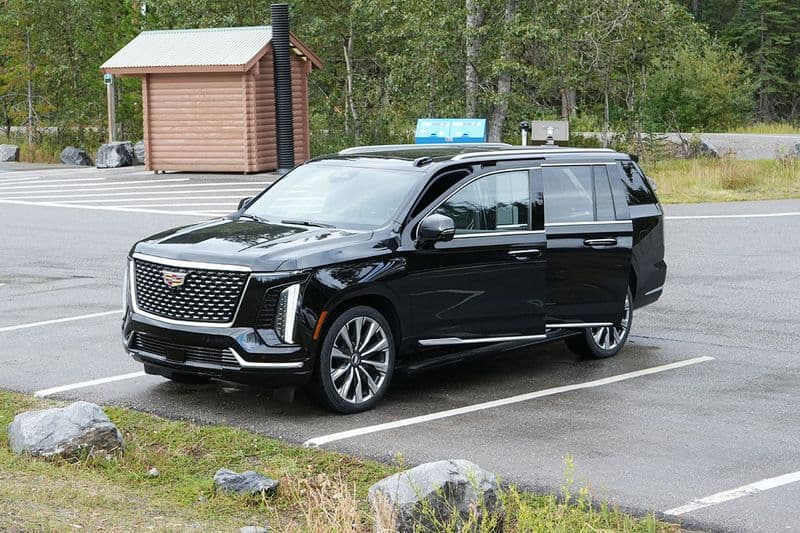 The Escalade parked at a mountain trailhead, seen from the front three-quarter with a log shelter and trees behind.