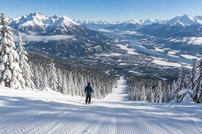 A skier at the top of a long groomed run above Revelstoke, the town and the Columbia River far below and snowy peaks all around.